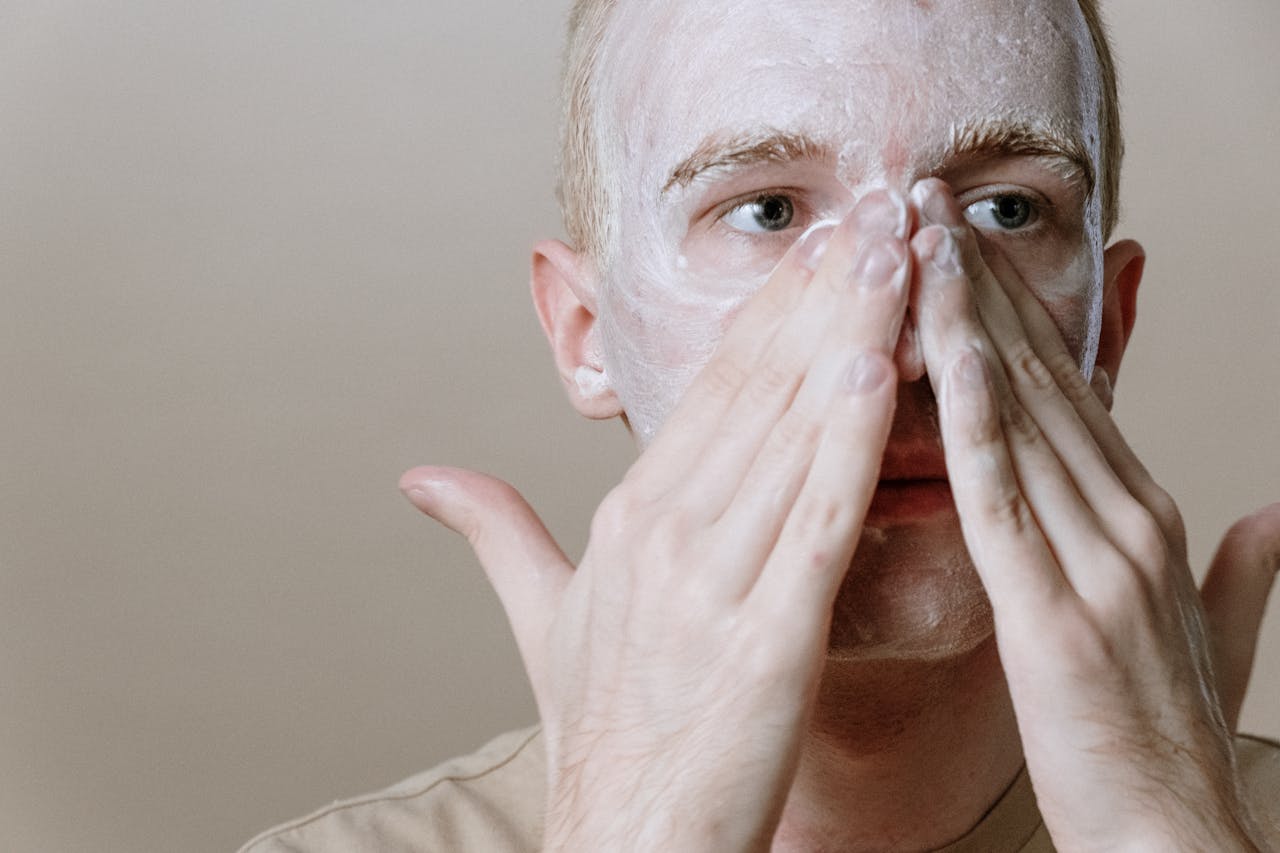 A young man applies skincare cream to his face, focusing on freshness and self-care in this indoor setting.